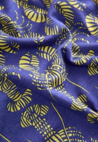 Tessuto blu con un motivo ripetuto di conchiglie nautilus gialle. La texture appare morbida e leggermente stropicciata, indicando un materiale leggero.