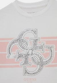 Valkoinen puuvillainen t-paita, jossa on vaaleanpunaiset raidat, suuri kimaltava hopeinen logo ja teksti "GUESS EST. 1981" alaosassa.