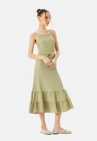 Abito maxi verde oliva, con texture, spalline regolabili, vita elastica e orlo con balza a strati. Abbinato a scarpe basse di tonalità chiara.