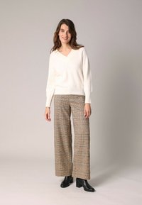 Femme portant un pull blanc à col V, un pantalon à carreaux marron à jambe large et des bottines à talons noirs, se tenant devant un fond uni.