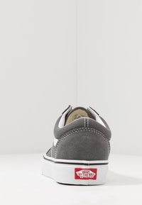 Baskets en toile suède grises avec accents blancs, dotées d'un profil bas et d'une semelle en caoutchouc. L'arrière présente une étiquette logo Vans rouge et une couture blanche.