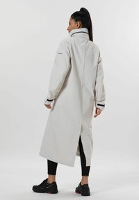 Lång, ljusgrå vattentät kappa med hög krage, svarta detaljer och en vent bak, parat med svarta leggings och skor.