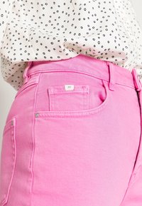 Vaqueros de mezclilla rosa de cintura alta, con dos bolsillos traseros, herrajes plateados y una etiqueta pequeña. Combinados con una blusa blanca con lunares.