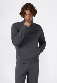 Giovane uomo con capelli ricci, che indossa una felpa zip-up Champion grigia scura e pantaloni abbinati, mano vicino al mento, su uno sfondo bianco semplice.