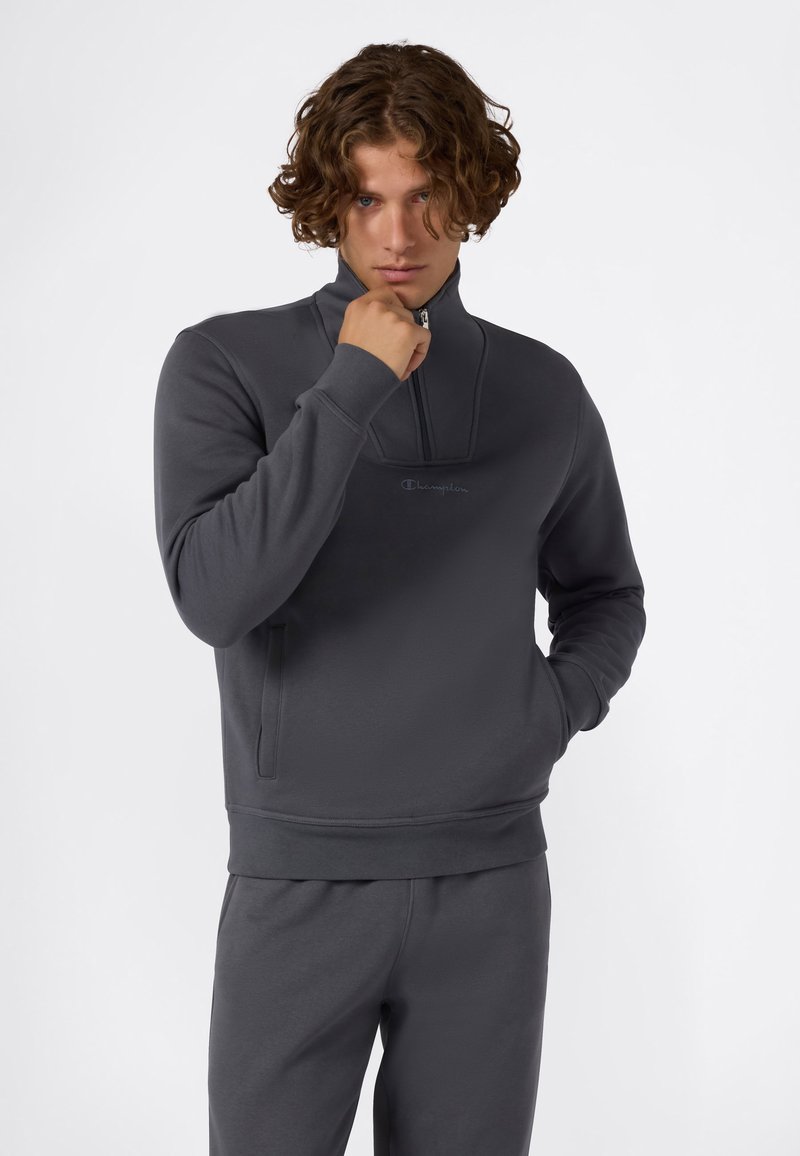 Giovane uomo con capelli ricci, che indossa una felpa zip-up Champion grigia scura e pantaloni abbinati, mano vicino al mento, su uno sfondo bianco semplice.