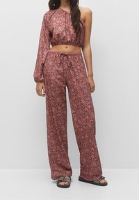 Femme portant un crop top asymétrique rouge à motifs avec une manche longue et un pantalon large assorti, sandales grises sur un fond uni.