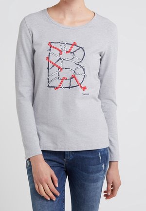 Jasnoszara koszula z długim rękawem z dużą literą „B” utworzoną z splecionych czerwonych i białych lin obramowanych granatowym kolorem, noszona przez osobę w niebieskich dżinsach.