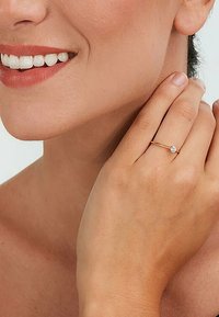 Golden Ring mit einem einzelnen klaren Edelstein, zarte Band, glatte Oberfläche und minimalistisches Design. Hand ruht am Hals und zeigt den Hautton.