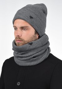 Pilkos spalvos megzta kepurė su sulenktu briaunotu kraštu ir trikampio formos logotipu, derinama su storu, briaunotu pilku šaliku, kuris apsuka kaklą.