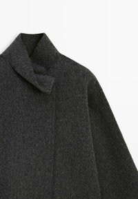 Tmavě šedý vlněný kabát s vysokým límcem a knoflíkovou stuhou na bílém pozadí, zobrazený v oblasti horního předního dílu a ramena.