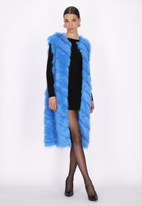 Blå faux fur-väst med en texturerad design och öppen front, bärs över en svart, figurnära klänning och parats med genomskinliga svarta strumpbyxor och klackar.