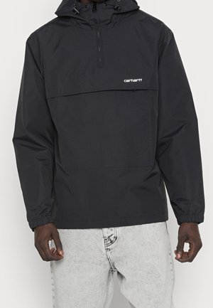 Veste à capuche noire Carhartt à enfiler avec poche avant et fermeture éclair, portée par une personne en jean clair, mains détendues sur les côtés.
