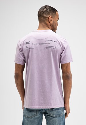 Mann trägt ein hellviolettes T-Shirt mit schwarzem Aufdruck am oberen Rücken und blaue Jeans, blickt von der Kamera weg.