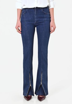 Femme portant un jean taille haute bleu foncé avec des fermetures éclair sur le devant à l'ourlet, associée à une chemise blanche et des chaussures pointues foncées.