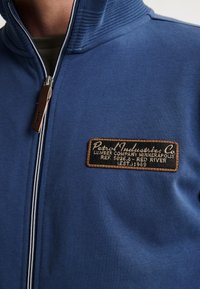 Giacca blu con zip e colletto a costine, caratterizzata da una patch in pelle marrone con testo. Realizzata in tessuto morbido, con dettagli in bianco sulla zip.