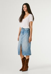 Jonge vrouw met lang donker haar, draagt een wit T-shirt met V-hals, een lichtblauwe denim rok tot op de knie met een voorste split, en bruine suède laarzen.