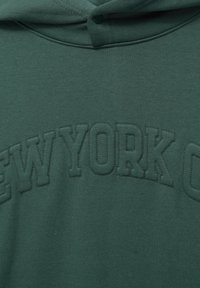 Sweat à capuche vert foncé avec le texte "NEW YORK" brodé sur la poitrine et un bouton-pression près de l'encolure de la capuche.