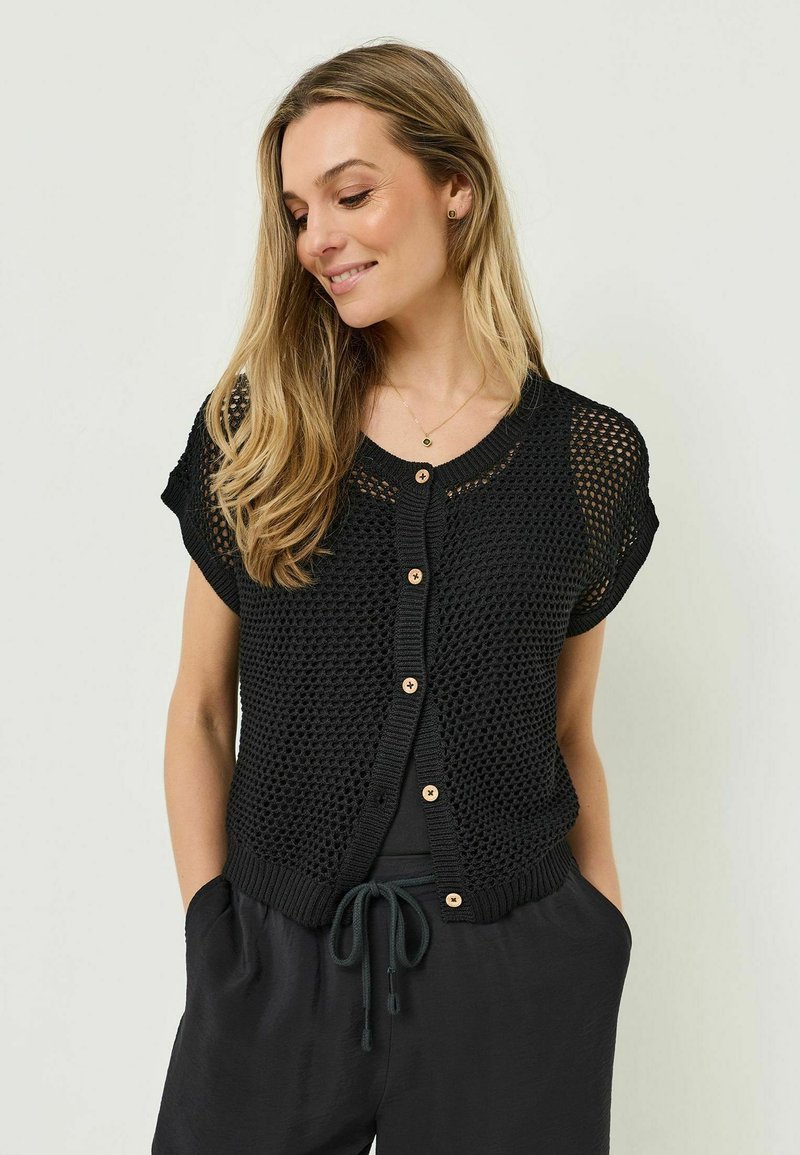 Femme aux longs cheveux blonds portant un cardigan noir à manches courtes en maille ajourée boutonné par-dessus un haut noir et un pantalon noir à cordon de serrage.