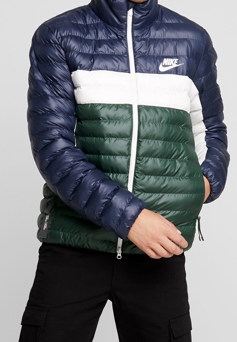 Vadderad jacka i marinblått, vitt och grönt. Har en hög krage, dragkedjeslutning och textilier med horisontella sömmar.