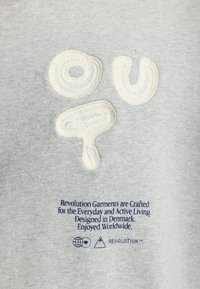 Grijze sweatshirt met textuur in crèmekleurige borduurwerk met de letters "ou" en een cirkelvormig ontwerp. Inclusief blauwe gedrukte tekst onderaan.