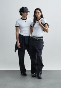 T-shirts brancas com costura canelada e acabamento preto, combinadas com calças pretas estampadas com logótipos. Acessórios incluem uma mala e um cinto.