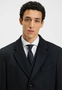Cappotto di lana nero con colletto a risvolto, chiusura con un solo bottone e taschino sul petto. Indossato su una camicia bianca e una cravatta blu navy.