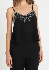 Haut sans manches noir avec un décolleté en V, orné de fil argenté et de perles en forme de feuilles le long du décolleté.