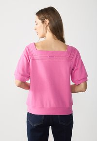 Felpa rosa con scollo quadrato, maniche corte a palloncino e dettagli traforati sulla parte superiore. Il tessuto sembra morbido con una finitura liscia.