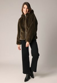 Veste à capuche en fausse fourrure khaki avec fermeture éclair à l'avant, présentant une texture douce et de grandes manches, associée à un pantalon large noir et des bottines.