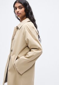 Μπεζ καμπαρντίνα, ελαφρύ υλικό, διπλό στήθος, μεγάλες τσέπες, άνετη εφαρμογή και κλασικό γιακά.