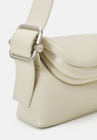 Borsa a tracolla in pelle beige con cima piegata, forma triangolare e accessori in metallo argentato sulla tracolla, che mette in risalto una superficie liscia.