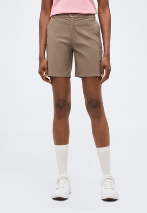 Person iført beige knælange shorts, hvide sokker, hvide sneakers og en lyserød skjorte, stående med hænderne afslappet ved siden.