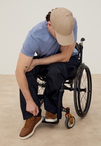 Man in wheelchair wearing a beige cap, blue shirt, and dark pants, tying the lace of his brown sneaker while seated indoors.