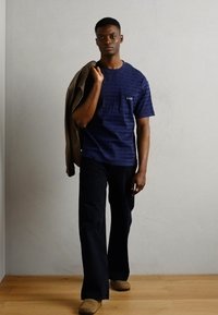 Chemise bleue marine à manches courtes avec des motifs texturés, associée à un pantalon sombre. Le mannequin porte une veste marron clair sur une épaule, se tenant sur un sol en bois.