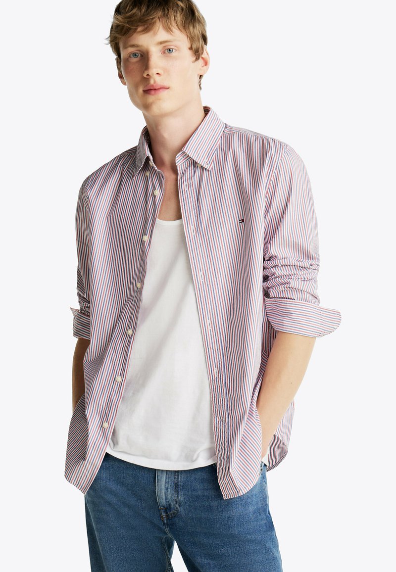Camisa de manga larga a rayas rojas y azules con cuello abotonado, botones blancos y un corte casual, llevada sobre una camiseta sin mangas blanca.