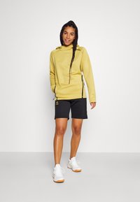 Sweatshirt à capuche jaune avec un détail de fermeture éclair, associé à un short noir. La tenue comprend des chaussures de sport blanches avec des accents orange.