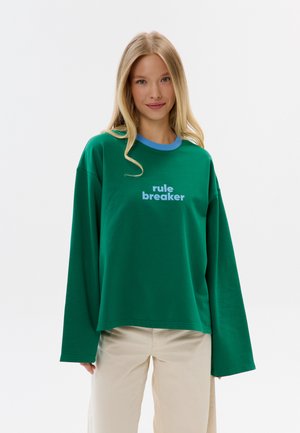 Jonge vrouw met lang blond haar die een groene oversized trui draagt met de tekst "rule breaker" en beige broek, staand op een witte achtergrond.