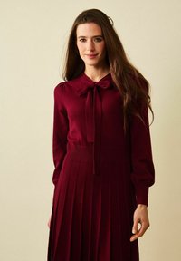 Burgundy-mekko, jossa on solmioneck, pallomaiset hihat ja pleatattu hame. Pehmeä kangas, jossa on ribattua vyötäröä. Rakenne näyttää tekstuurilta.