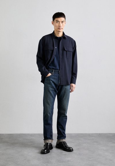 Marineblaues Hemd mit Knöpfen und zwei Fronttaschen, kombiniert mit dunkelblauen Jeans und schwarzen Lederschuhen. Geschmeidige Textur, lässige Passform und hochgekrempelte Ärmel.