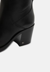 Botine negre din piele cu toc bloc înălțat. Prezintă o textură netedă, cusături subtile și un accent metalic cu logo pe toc.