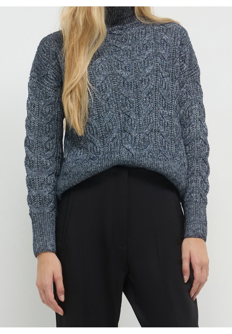Femme portant un pull à col roulé gris foncé en maille torsadée et un pantalon noir taille haute, avec de longs cheveux blonds sur une épaule.