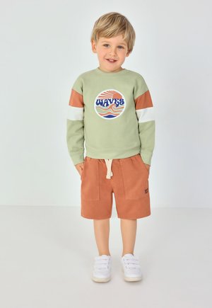 Zelená mikina s oranžovými a bílými pruhovanými rukávy, s kulatým grafikou "WAVES"; v kombinaci s oranžovými šortkami a bílými teniskami.