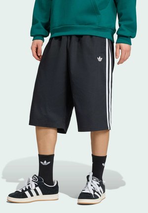 Man die zwarte Adidas-shorts met witte strepen draagt, zwarte Adidas-sokken en zwarte Adidas-sneakers, staand tegen een effen achtergrond.