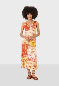 Maxi-Kleid in Creme mit roten und orangefarbenen Tropenmotiven. V-Ausschnitt-Design, ärmellos, mit einem gemusterten Textil. Getragen mit braunen Sandalen.
