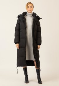 Frau in einem langen schwarzen gepolsterten Mantel mit Kapuze über einem grauen gerippten Rollkragenkleid und schwarzen kniehohen Stiefeln, stehend vor einem schlichten Hintergrund.