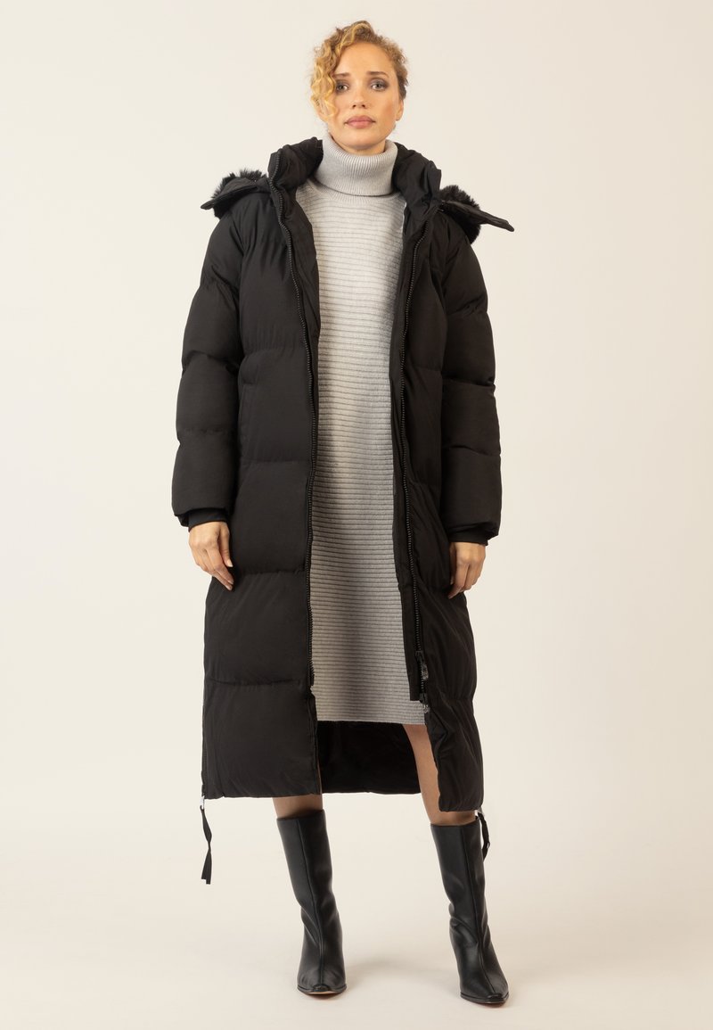 Frau in einem langen schwarzen gepolsterten Mantel mit Kapuze über einem grauen gerippten Rollkragenkleid und schwarzen kniehohen Stiefeln, stehend vor einem schlichten Hintergrund.