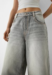 Mujer vestida con jeans de pierna ancha de lavado claro y una camiseta corta blanca, de pie contra un fondo claro y liso.