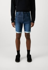 Șorturi din denim cu spălare în albastru închis, cu margine rulată, design cu cinci buzunare și estompare subtilă pe partea din față, asortate cu un hanorac negru.