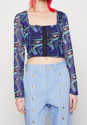 Person wearing a colorful, abstract-print long-sleeve crop top with black button details, paired with light blue pants featuring gold buttons.