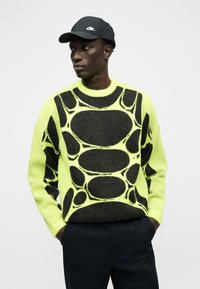 Άνδρας που φοράει μαύρο καπέλο Nike, φωσφορίζουσα κίτρινη μπλούζα με μεγάλα μαύρα αφηρημένα σχήματα και μαύρο παντελόνι, στέκεται με τα χέρια στις τσέπες.